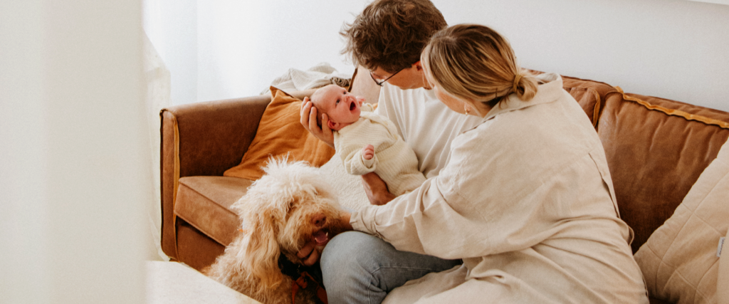 Μια τριμελής οικογένεια με ένα μικρό σκυλάκι σε έναν άνετο καφέ καναπέ, απολαμβάνουν μια χαρούμενη στιγμή με το μωρό τους.